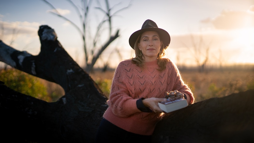 A woman leans against a fallen tree against the sunset.  She is wearing a hat and is holding a book.