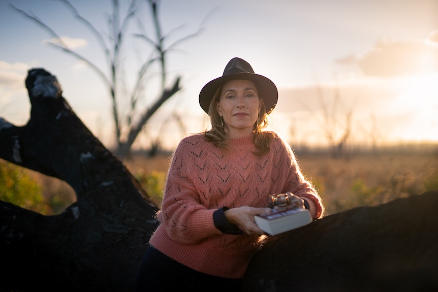 A woman leans against a fallen tree against the sunset. She's wearing a hat and holding a book.