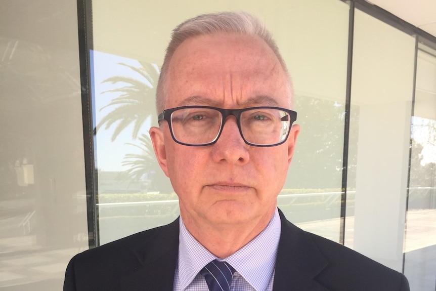 A man in a suit and wearing glasses stands in front of the window of a building.