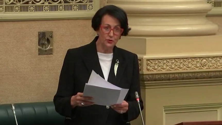 A woman with black hair wearing a black suit and leaf brooch holds a piece of paper next to a large column