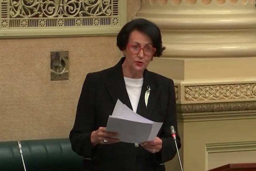 A woman with black hair wearing a black suit and leaf brooch holds a piece of paper next to a large column