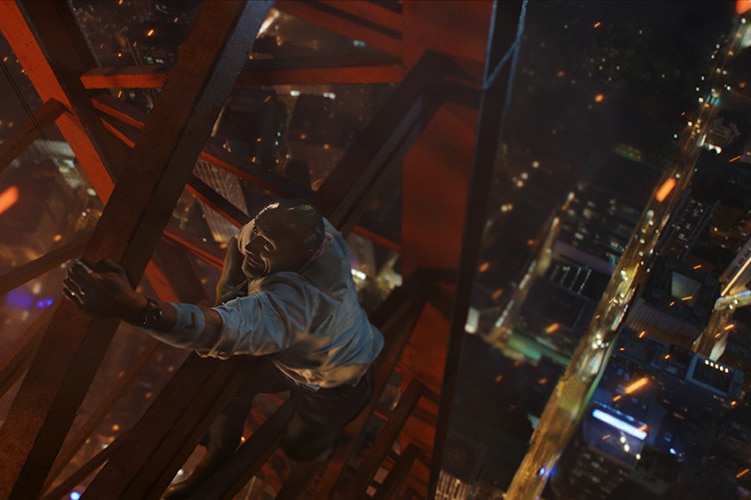 Still image from 2018 film Skyscraper of Dwayne Johnson hanging from a construction structure high above a cityscape.