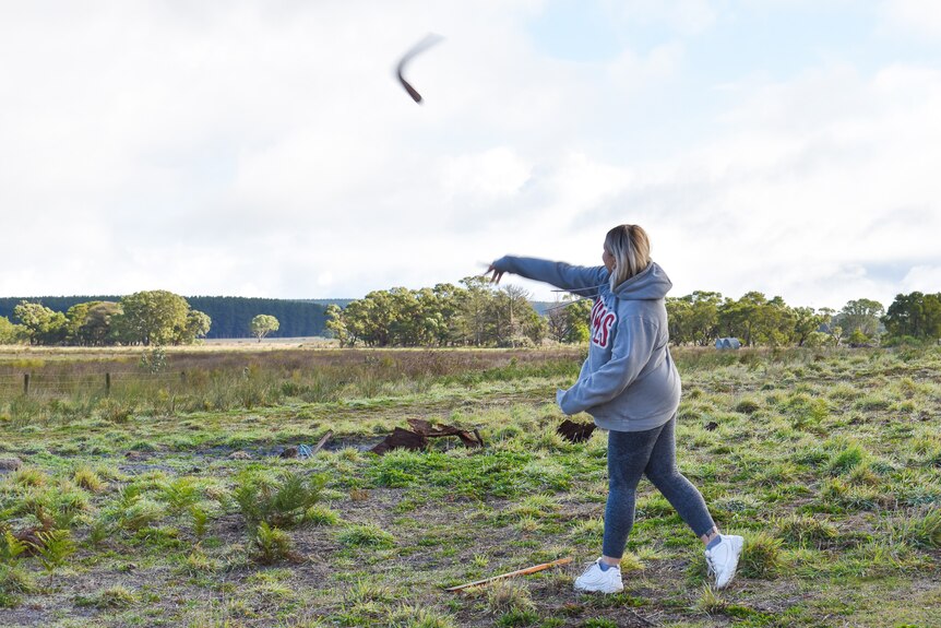 A woman with blonde hair in a grey houded jumper throws a boomerang into the distance, forest trees behind her.