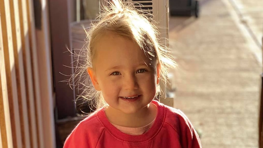 Cleo Smith wearing a red jumper standing outside and smiling for a photo.