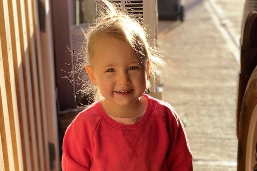 Cleo Smith wearing a red jumper standing outside and smiling for a photo.