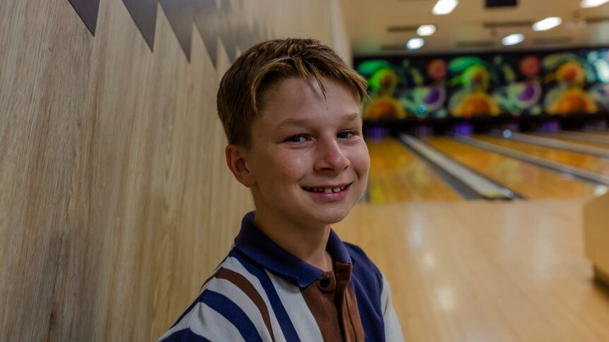 A young boy sits and smiles at the camera. Bowling alley in background.