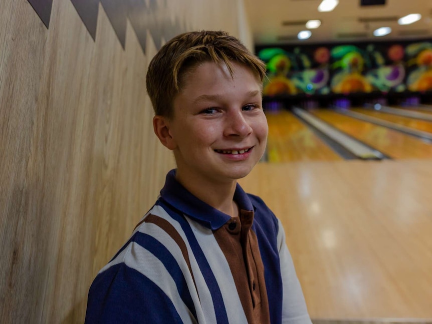 A young boy sits and smiles at the camera. Bowling alley in background.