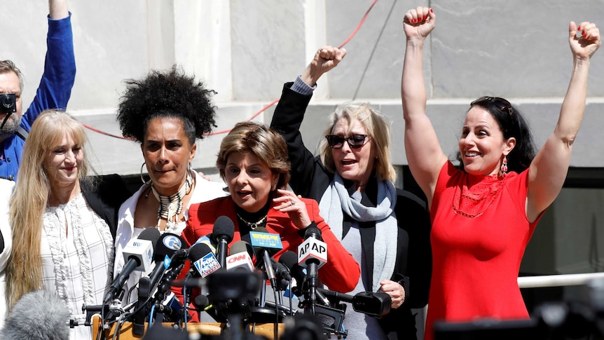 Accusers, some with their hands in the air, stand behind a lawyer holding a press conference.
