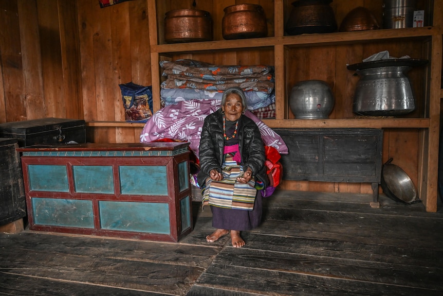 A woman sits on a chair in traditional clothes with a black puffer jacket on top. huge copper containers sit on a shelf behind 