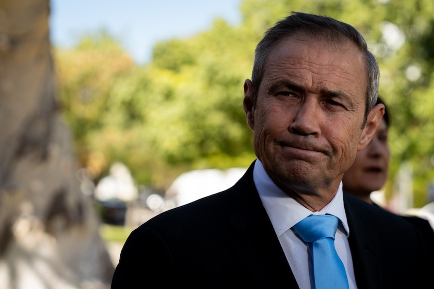 Roger Cook in a black jacket and blue tie, pressing his lips together.