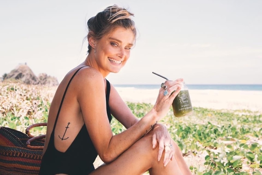 A woman sits on a beach holding a green juice