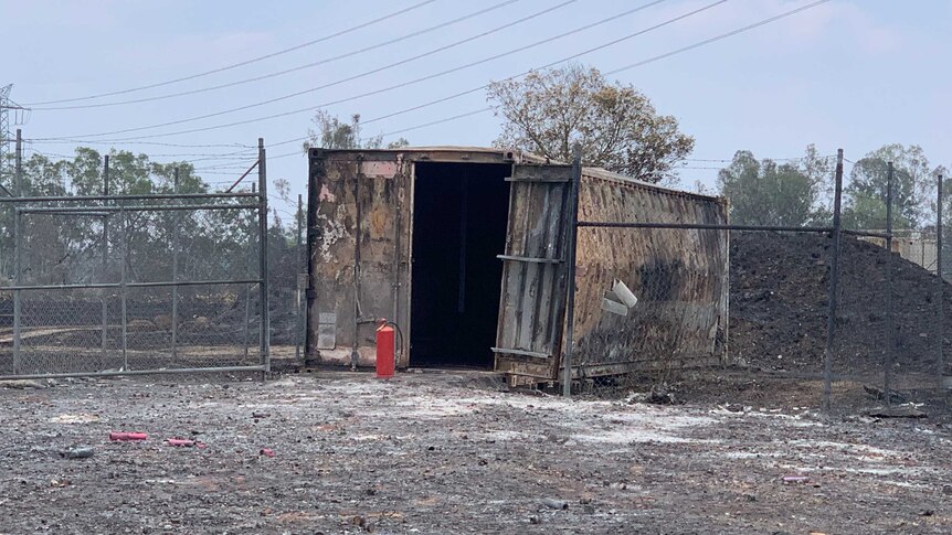 A shipping container that is burnt and one side is warped. One of it's doors is ajar and the ground is blackened.