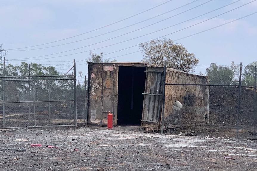 A shipping container that is burnt and one side is warped. One of it's doors is ajar and the ground is blackened.