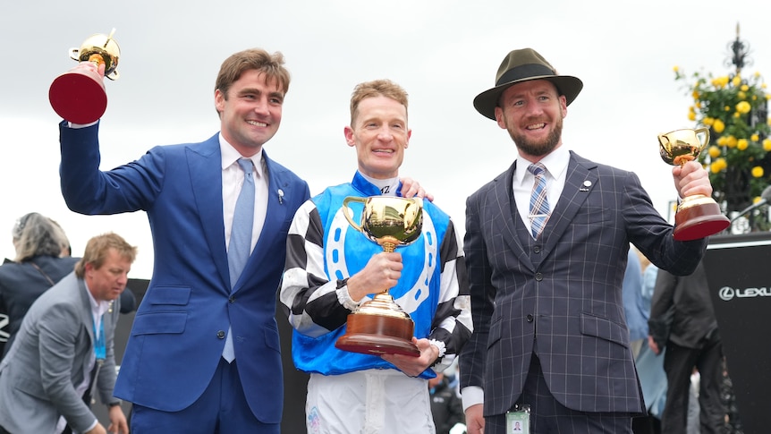 The two trainers of the Melbourne Cup winner stand either side of the winning jockey, all holding trophies after the race. 