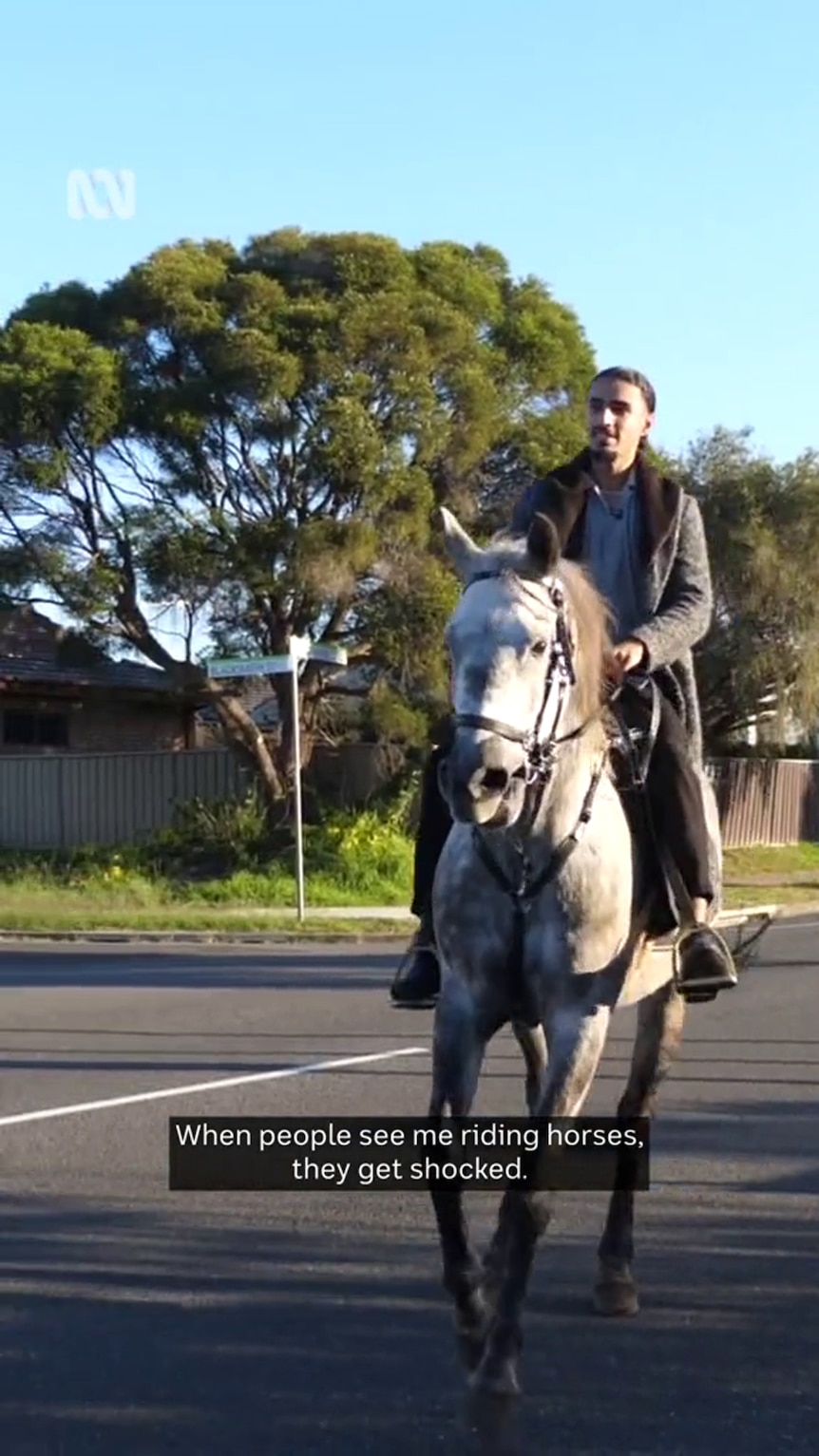 A young man with a dark beard and medium-tone skin rides a black and white horse on a suburban street.