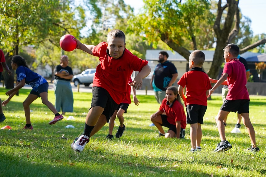 A young boy in the foreground running while holding a dodge ball. Several other kids in the background. 