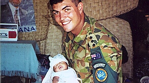 Ben in army fatigues kneeling down in the camp holding baby ben in his arms