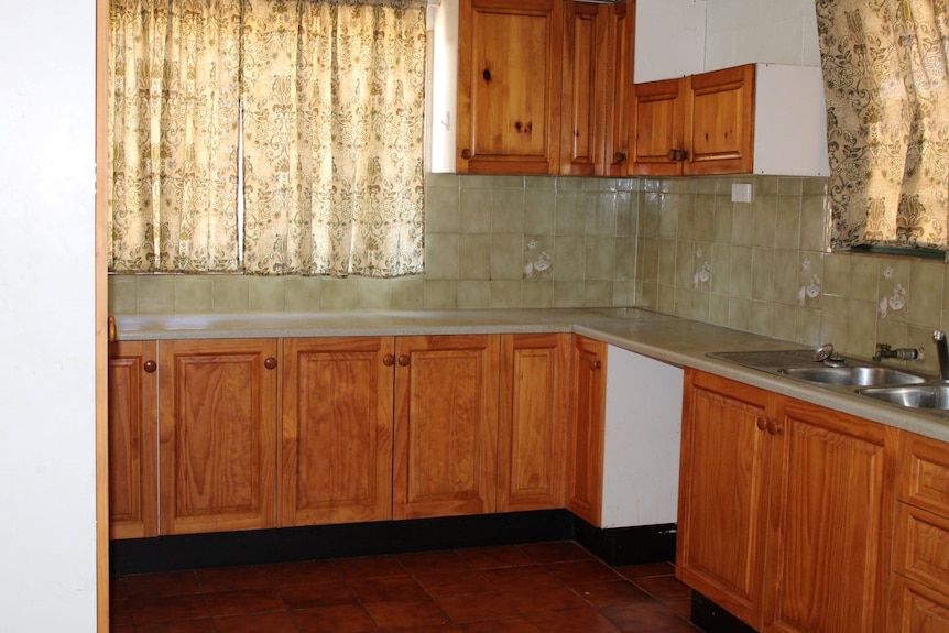 A green tiled kitchen with wooden cabinets and a hole where there should be a stove, printed curtains.