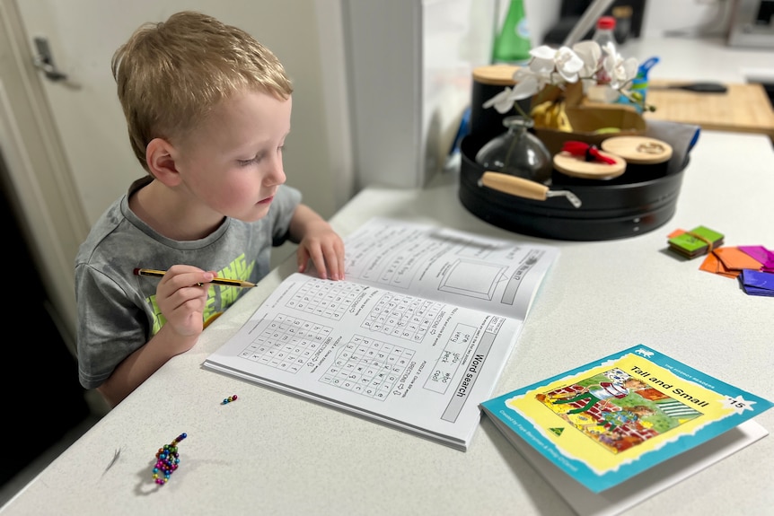 A young boy sits at a table with a pencil, looking a book of writing exercises.