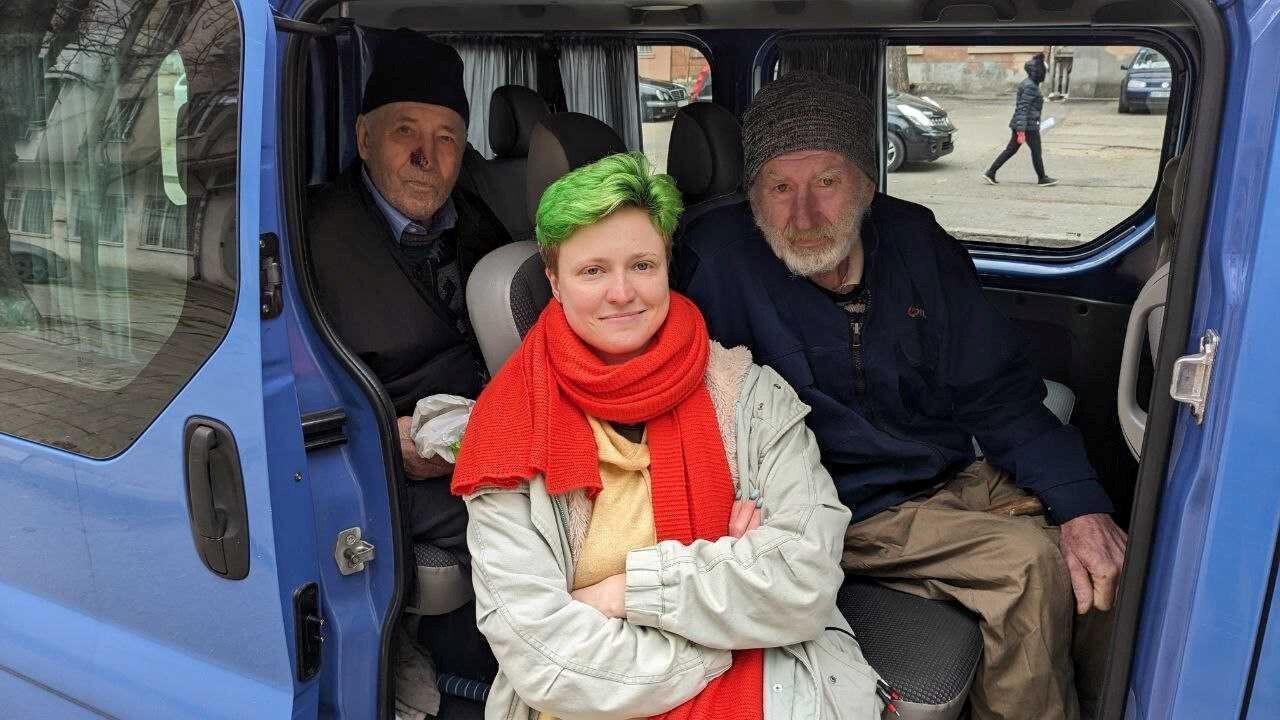 A young woman with green hair stands at the open door of a mini-van posing with two elderly men in the van behind her. 