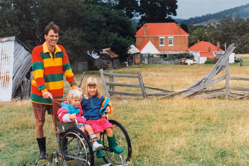 A man pushing two small daughters along in a wheelchair in the country
