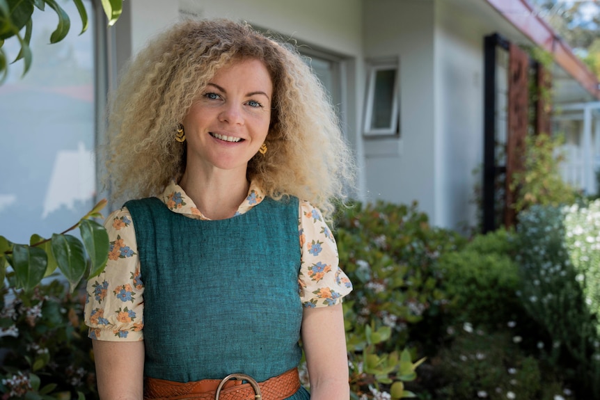 Woman with blonde curly hair wearing a floral yellow and green dress standing outside.