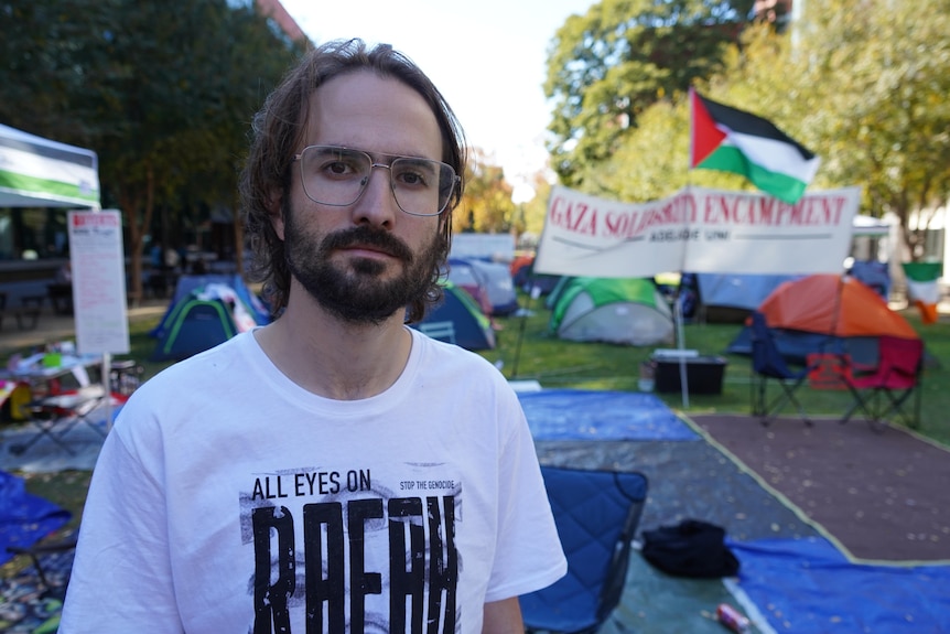 A man in a white t-shirt stands in front of a group of tents.