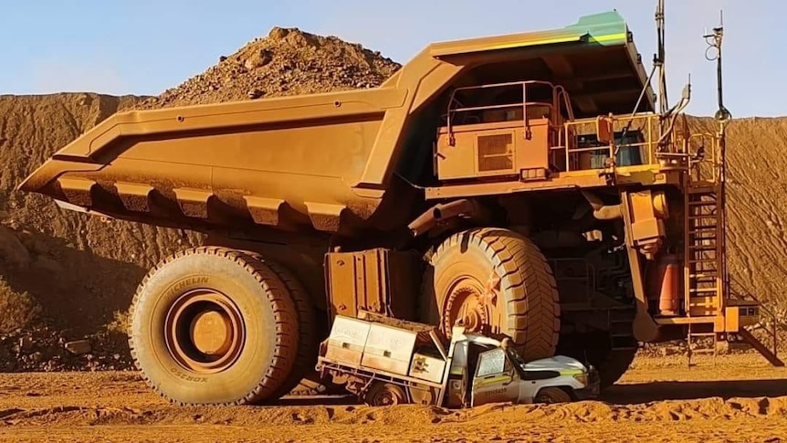 A white four wheel drive ute is crushed beneath the tyre of a large yellow dump truck