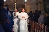 a young man in a white robe leads a procession of priests through a Catholic church