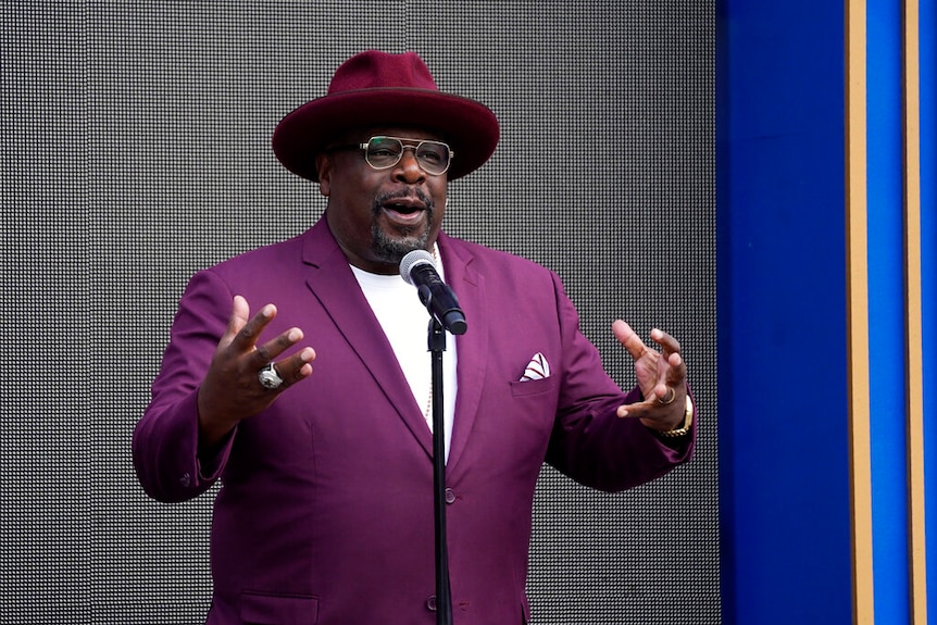 Cedric the Entertainer in a colourful suit, speaking at a microphone.