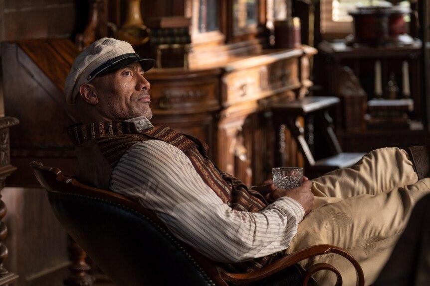 Dwayne Johnson leans back in an old chair with a firm expression, holding a glass of brown spirits in his hand
