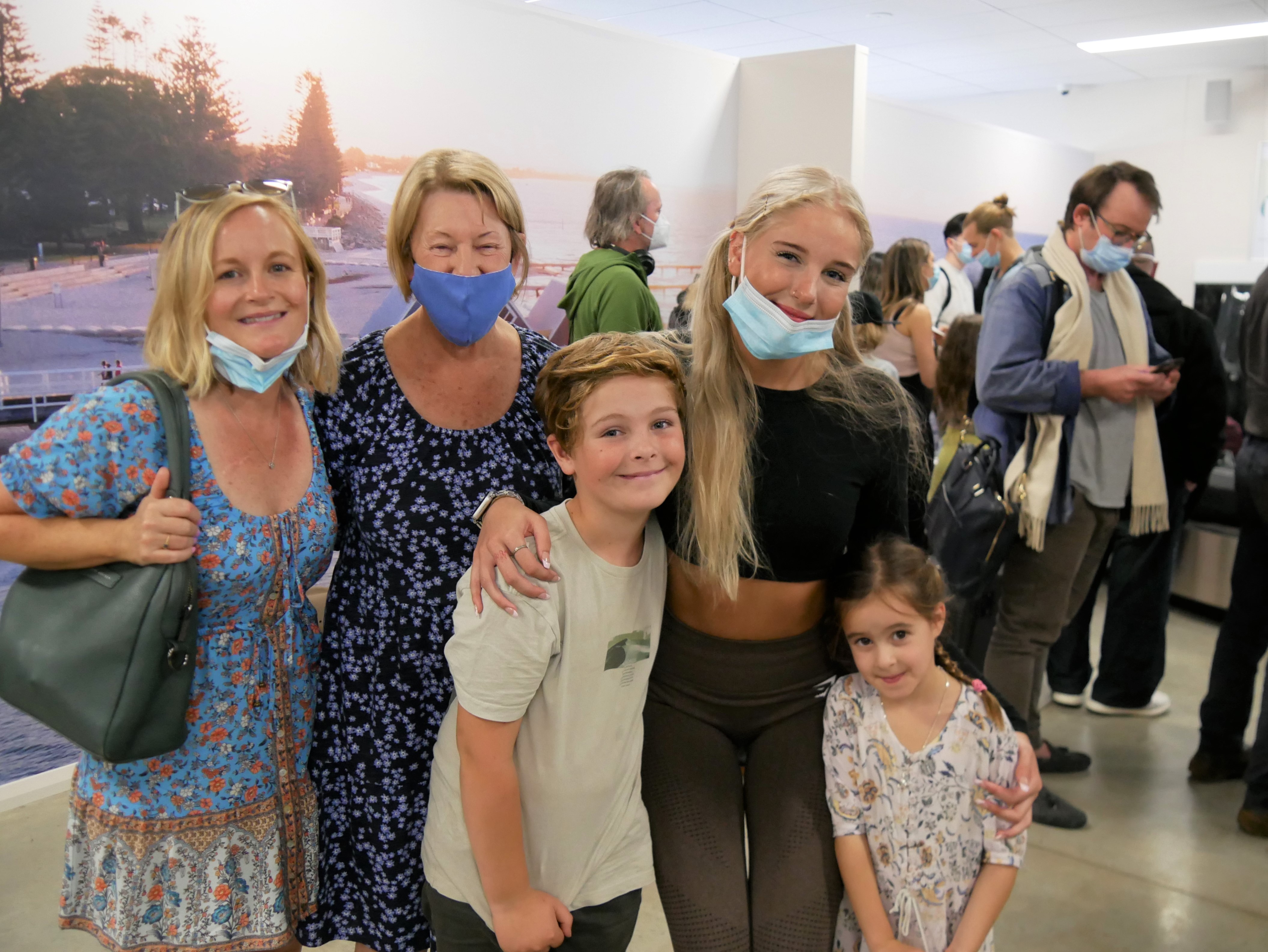 Three women, a young boy and girl, all embrace each other and smile at the arrivals terminal. 