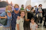 Three women, a young boy and girl, all embrace each other and smile at the arrivals terminal. 