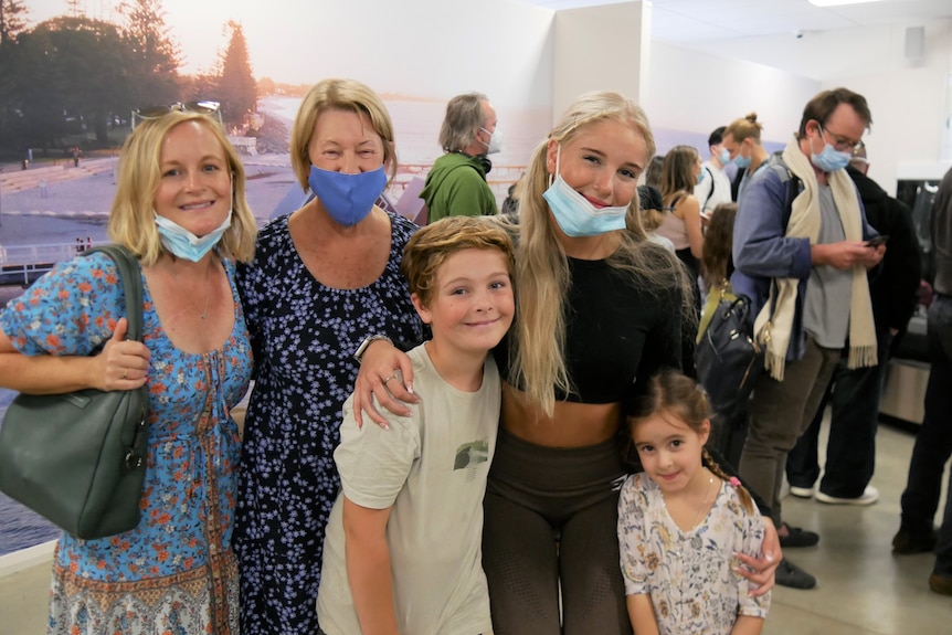 Three women, a young boy and girl, all embrace each other and smile at the arrivals terminal. 