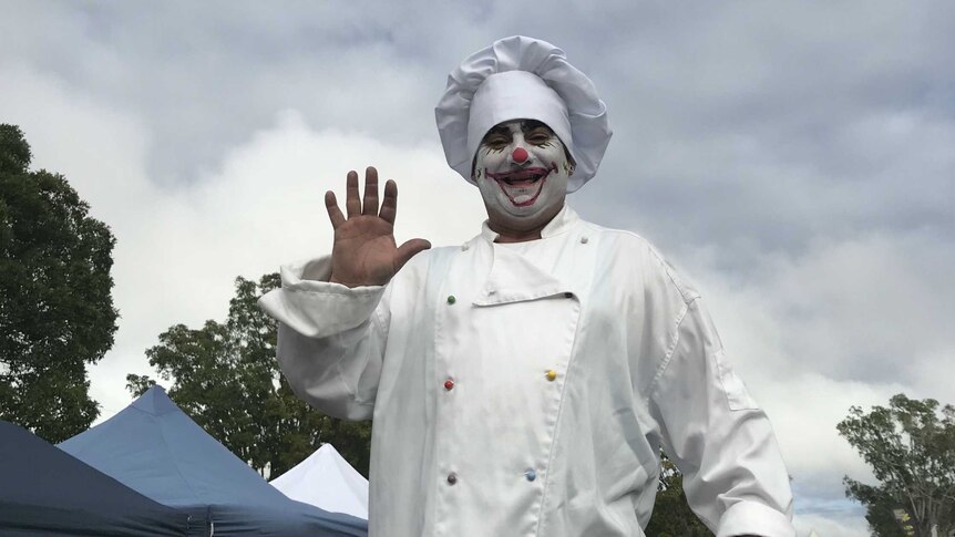 Chef with face painted as a clown stands smiling with his hand raised, in front of a cloudy sky.