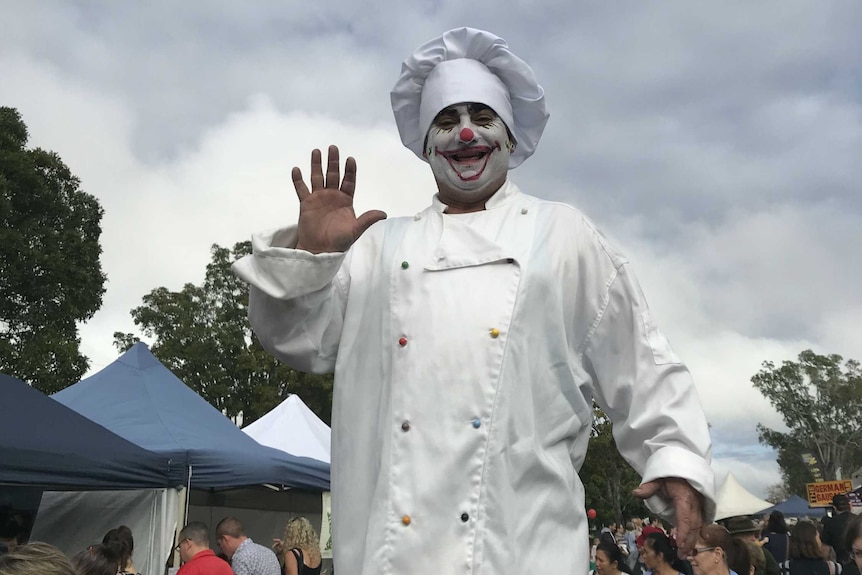 Chef with face painted as a clown stands smiling with his hand raised, in front of a cloudy sky.