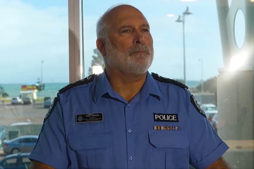 A man with a beard stands in a blue police uniform, in front of a window