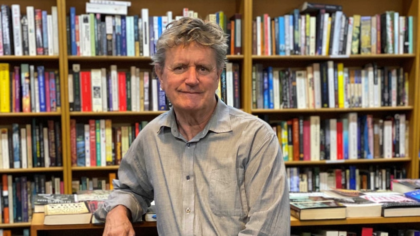 A man holding a book poses in front of a bookshelf in a shop