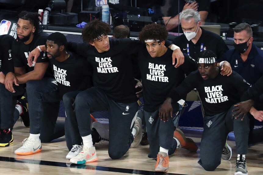 New Orleans Pelicans players kneel on the side of the court, arm in arm. They are wearing shirts that read "BLACK LIVES MATTER"