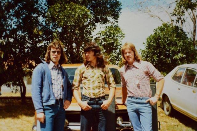Old photo of three young men with long hair standing in front of an orange car.