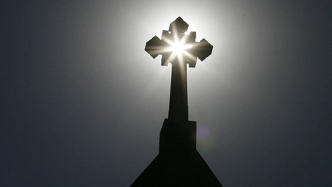 A church's cross is silhouetted against the sun and a grey sky.