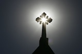 A church's cross is silhouetted against the sun and a grey sky