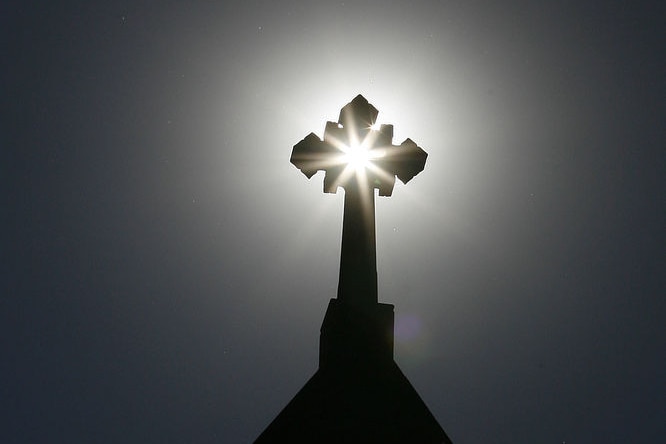 A church's cross is silhouetted against the sun and a grey sky