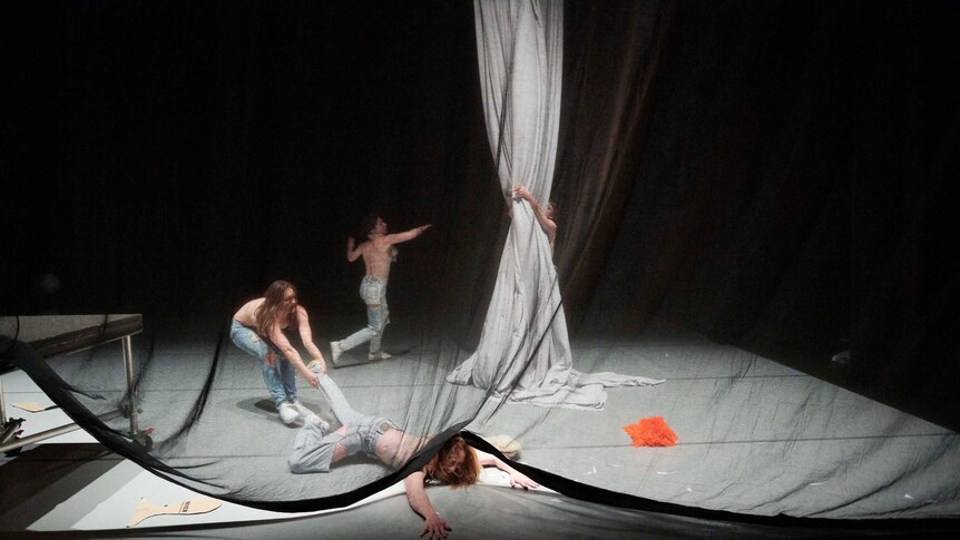 A square stage with dancers, 1 dragging another on the ground, 1 in background dancing frenetically, 1 grabbing white material