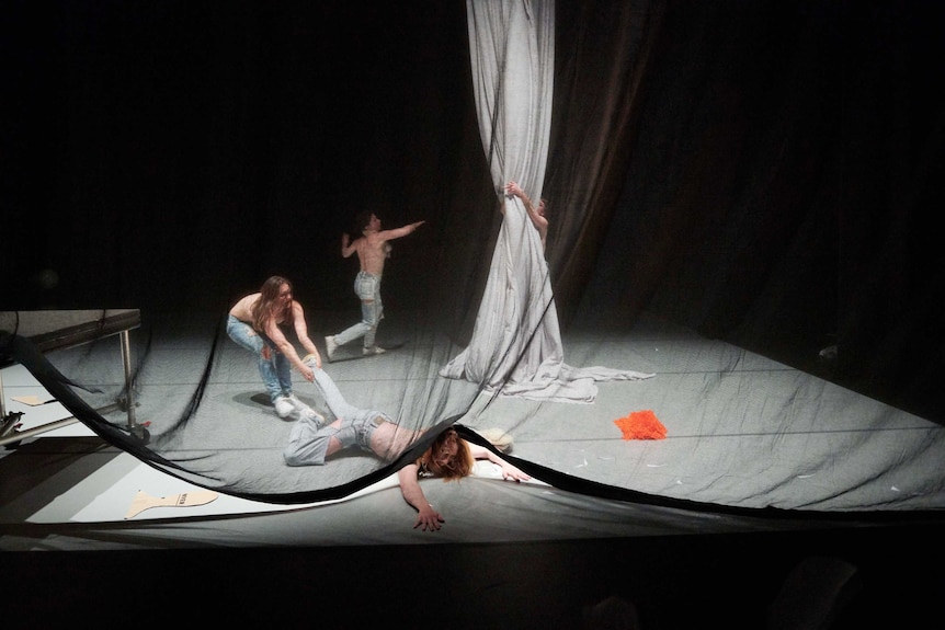 A square stage with dancers, 1 dragging another on the ground, 1 in background dancing frenetically, 1 grabbing white material