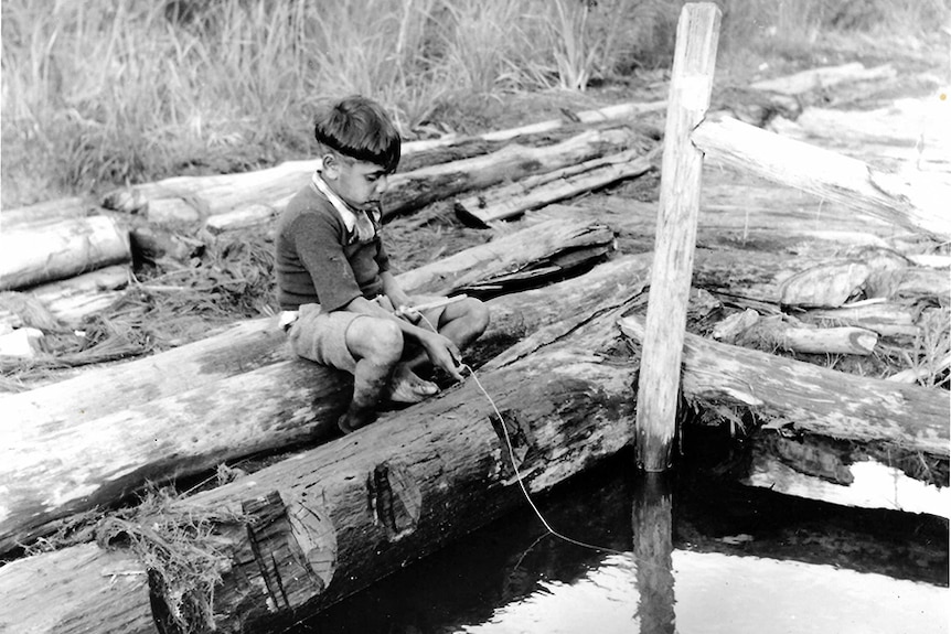 A young Indigenous boy sits on a log fishing in the creek 