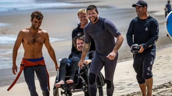 Barney Miller being pushed up the beach in a wheelchair by his mates