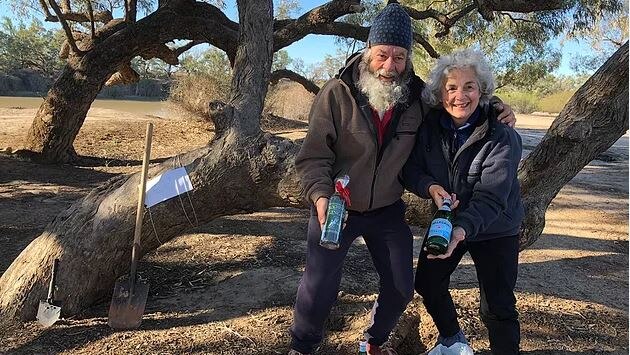 Phil and Susan McDonald pose with bottles of mineral water in front of a tree with a shovel lying against it.