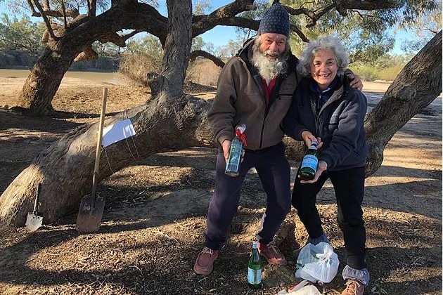 Phil and Susan McDonald pose with bottles of mineral water in front of a tree with a shovel lying against it.