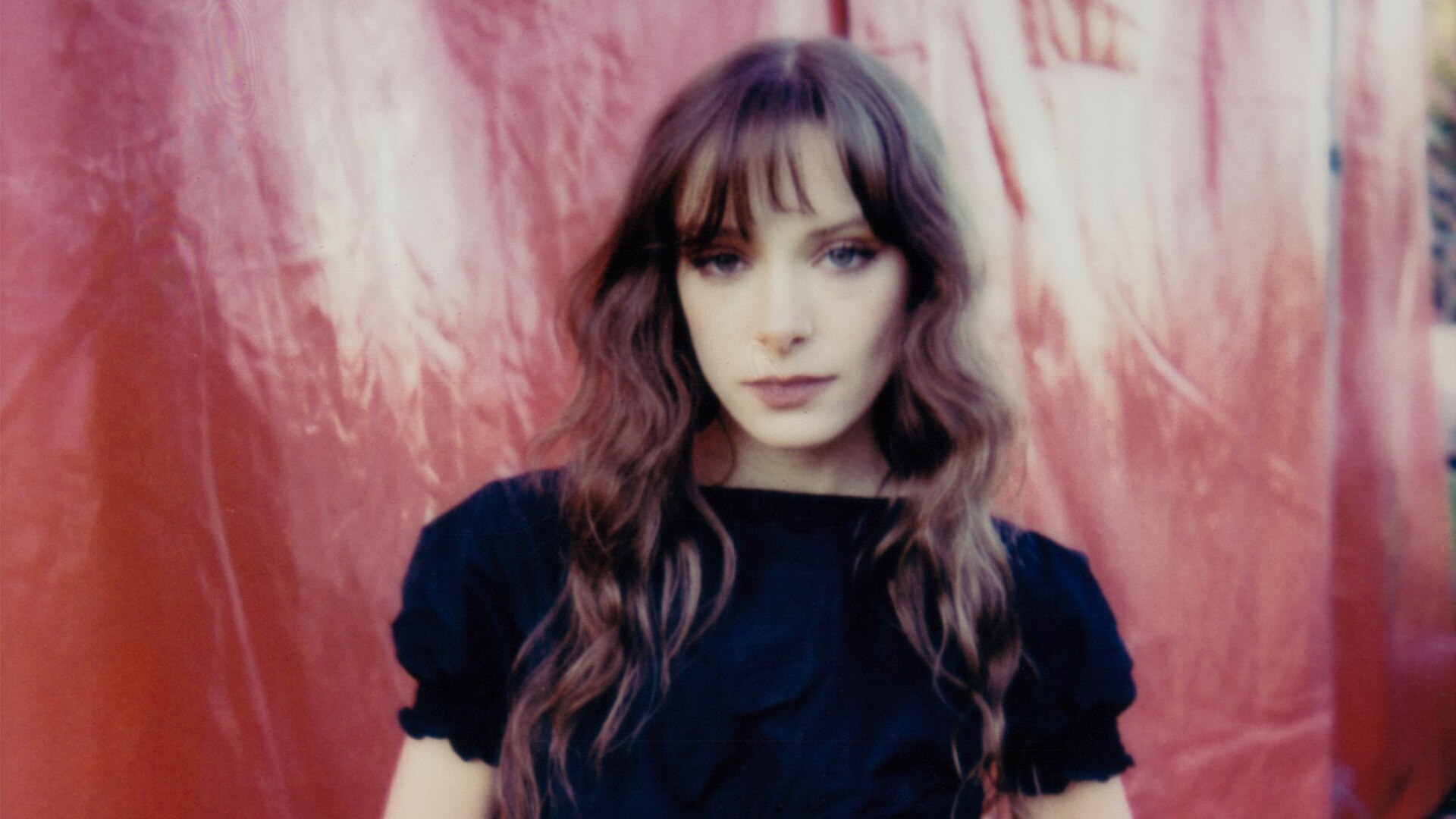 A woman with a fringe and brown, flowing hair stands in front of a red background. The photo is taken on film; the colours glow.